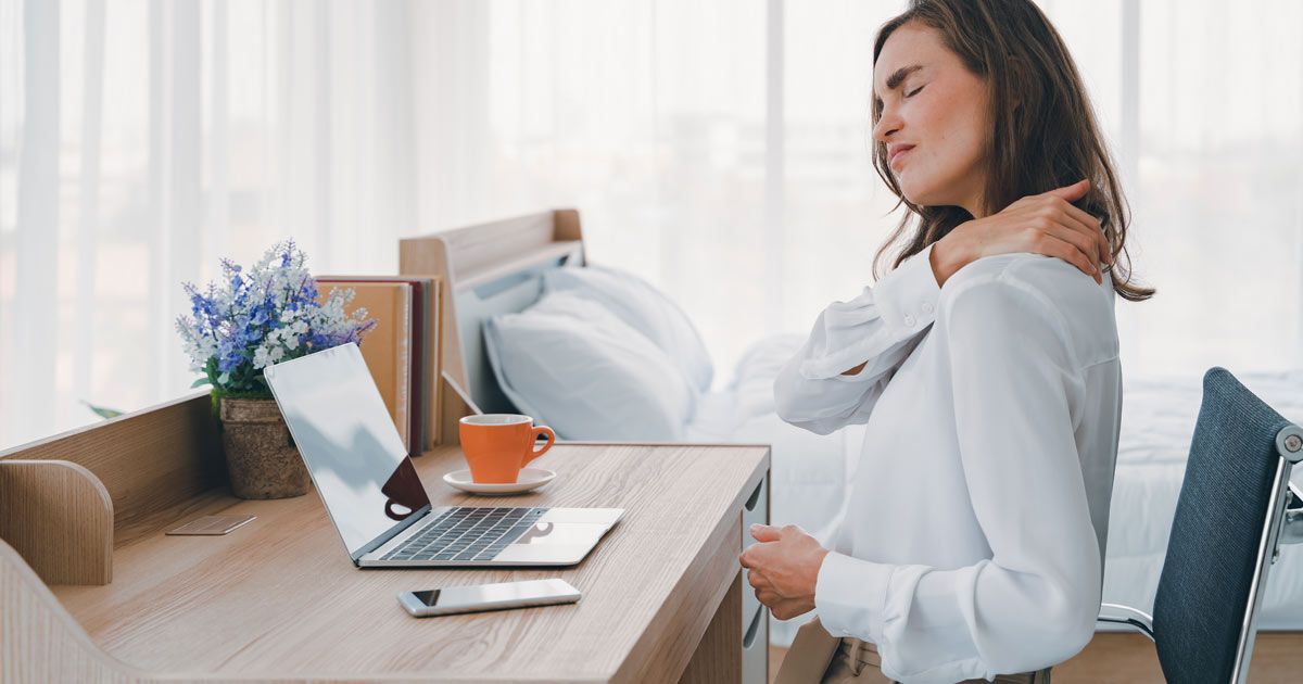 A woman at a desk, grimacing while holding her shoulder, suggesting pain. Laptop, coffee cup, and window in the background needing Fibromyalgia Treatments NYC - Dr. John Salerno