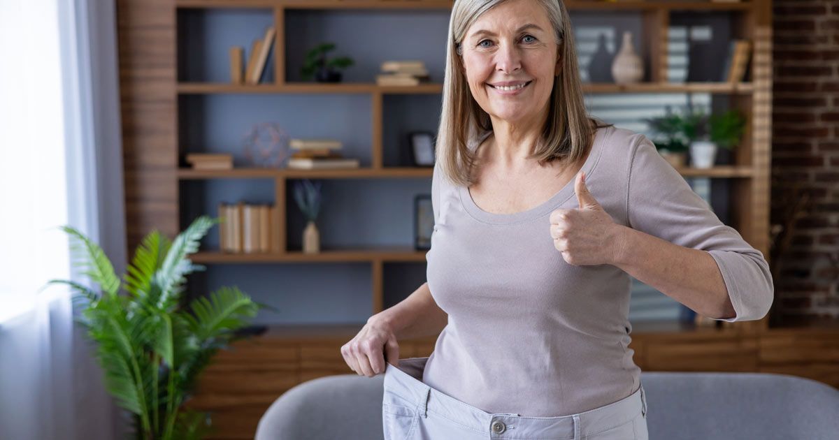 An older woman is standing in a living room holding her old pants and giving a thumbs-up for losing weight with GLP-1 Therapy NYC by Dr. John Salerno
