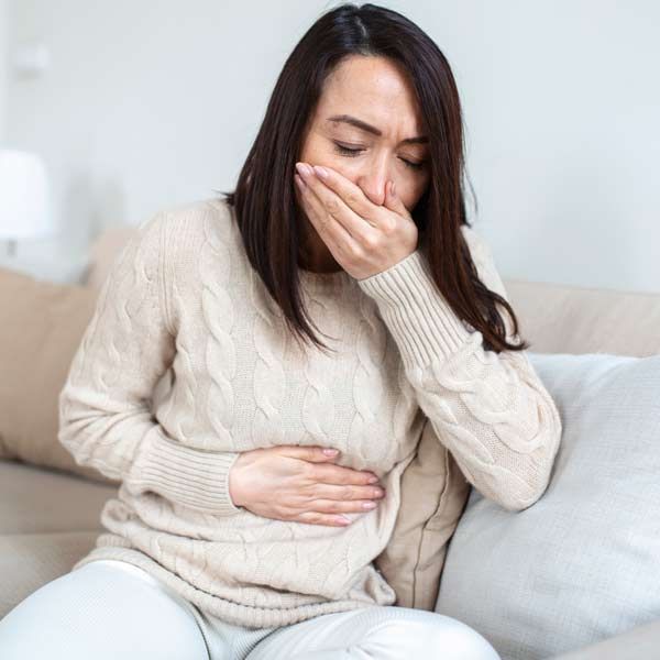 Nausea and stomach pain are common symptoms of Gastritis. A woman is sitting on a couch, covering her mouth with her hand, needing Gastritis Treatments