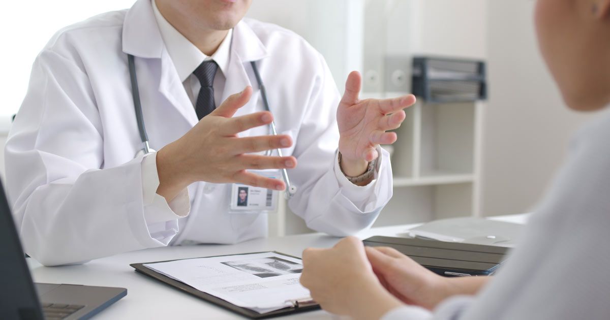 Doctor in white coat gestures with hands, speaking to a patient in a doctor's office at their Holistic Physical NYC by Dr. John Salerno