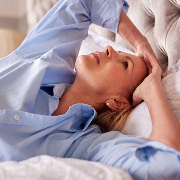Woman with blonde hair lying in bed, eyes closed, hand on her forehead, wearing a blue button-down shirt. She can't sleep and is need of HRT Treatments by Salerno Wellness in Manhattan, NY and Connecticut