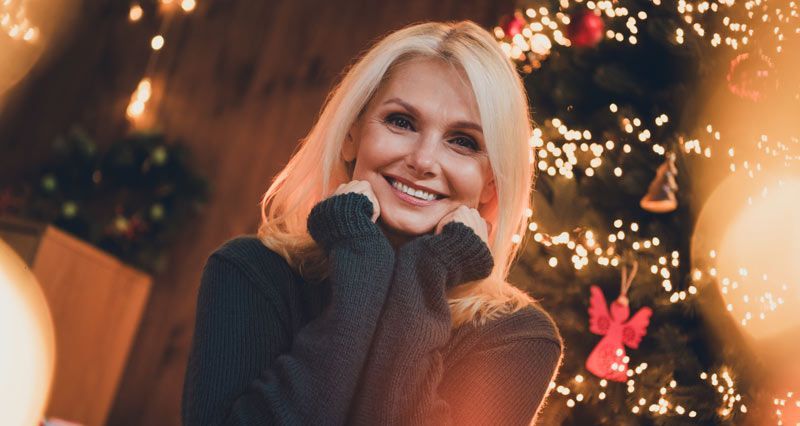 Woman smiling, posing in front of a Christmas tree after learning how to make her skin glow.