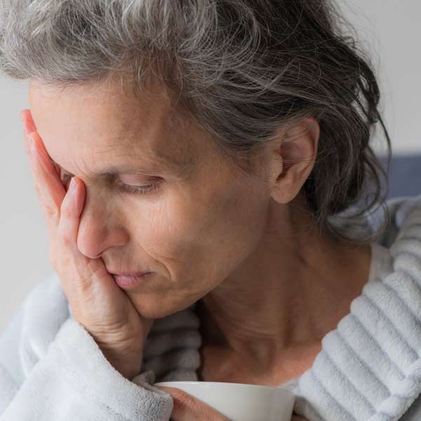 A woman with a weakened Immune System, gray hair, holds a cup, resting her hand on her face, appearing weary.