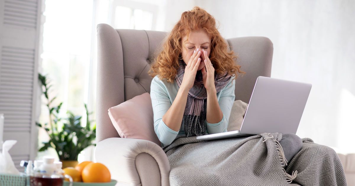 Woman with red hair, blowing nose, sitting in an armchair with a laptop, blanket, and tissues; appears ill needing Immunodeficiency Treatments NYC - Dr. John Salerno
