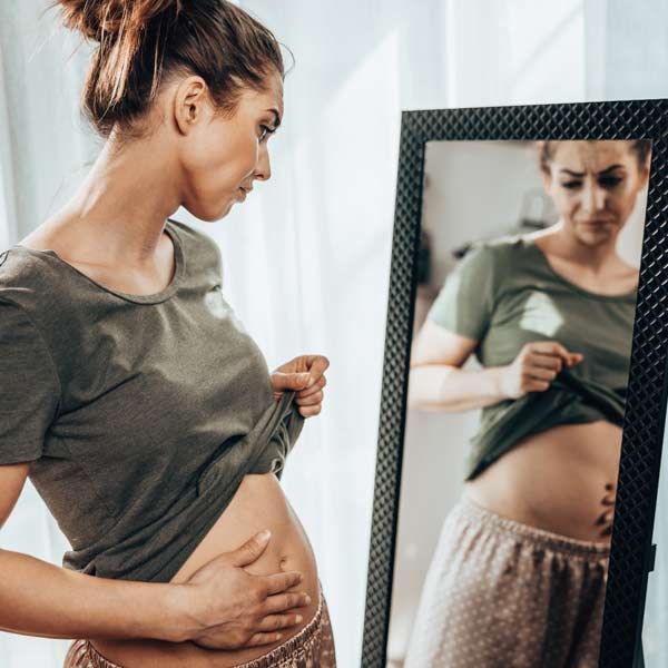 Bloating is an early indication of IBS. A woman is looking at her stomach in a mirror for Irritable Bowel Syndrome Treatments