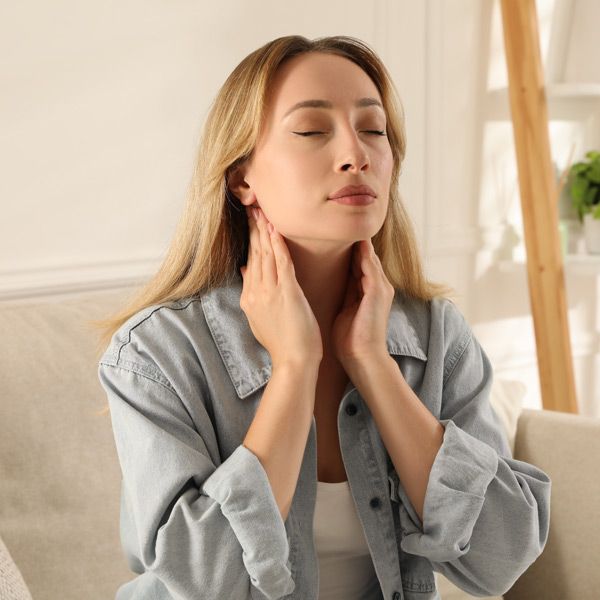 A woman is sitting on a couch with her eyes closed, touching her neck, representing Thyroid Disease Care