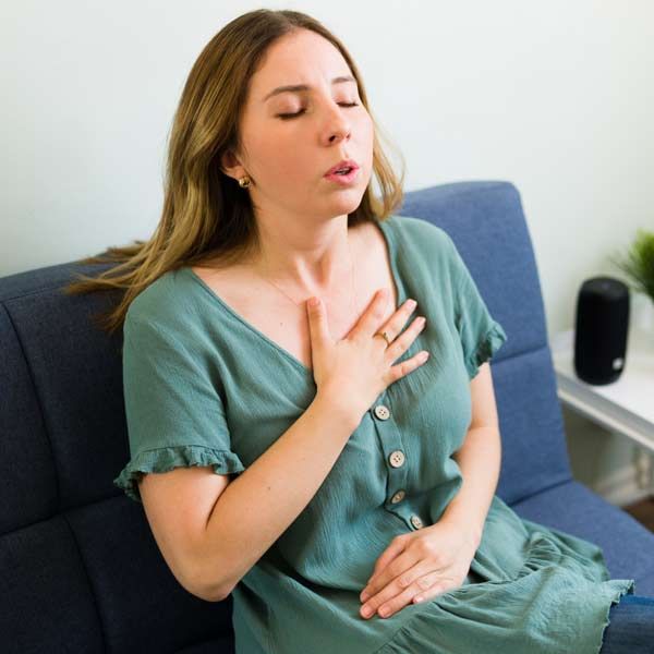 Woman sitting on a couch with eyes closed, hand on chest, couhing, a Candidates for Mold Poisoning Treatments by Dr. John Salerno