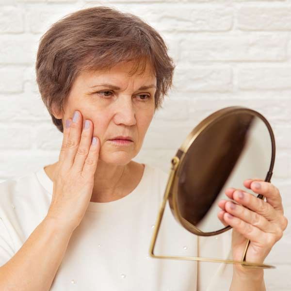 Woman looking at a mirror, touching her cheek with a concerned expression. White brick wall background. An Ideal Candidate for NAD+ Treatments NYC and CT