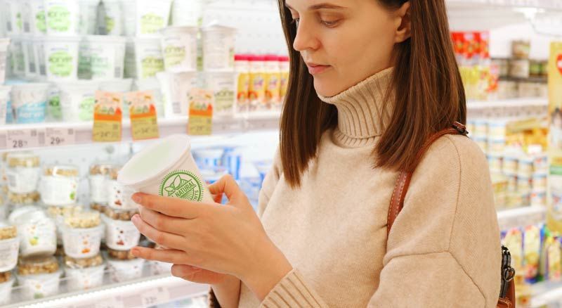 Woman in cream sweater reading label on yogurt container in a supermarket knowing Natural Foods Can Harbor Hidden Toxins