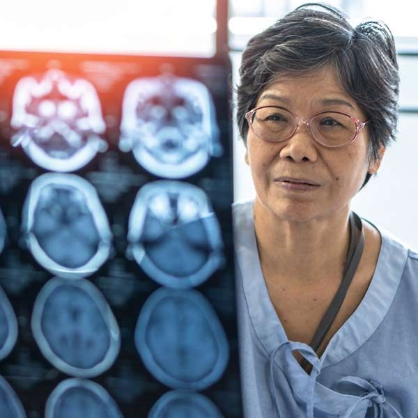 A woman is standing in front of an x-ray of her brain representing Neurodegenerative Conditions Treatments