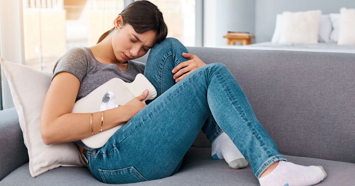 A woman is sitting on a couch holding a hot water bottle after scheduling Polycystic Ovary Syndrome Treatments NYC with Dr. John Salerno