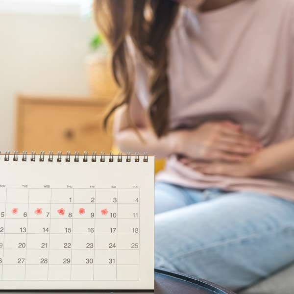 A woman is sitting on a couch holding her stomach next to a calendar representing Premenstrual Syndrome Treatments