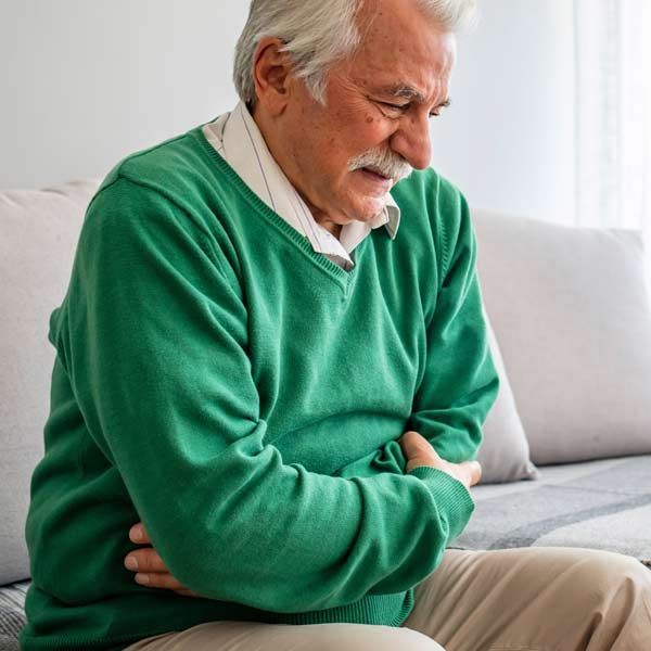 A man in a green sweater is sitting on a couch holding his stomach, seeking Complementary and Functional Medical Treatments for Natural Ulcerative Colitis Relief by Dr. John Salerno in Manhattan, NY, and Connecticut