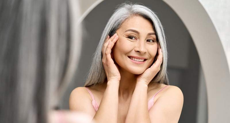 Woman with gray hair smiling in mirror, hands on her face; light pink top; neutral background.