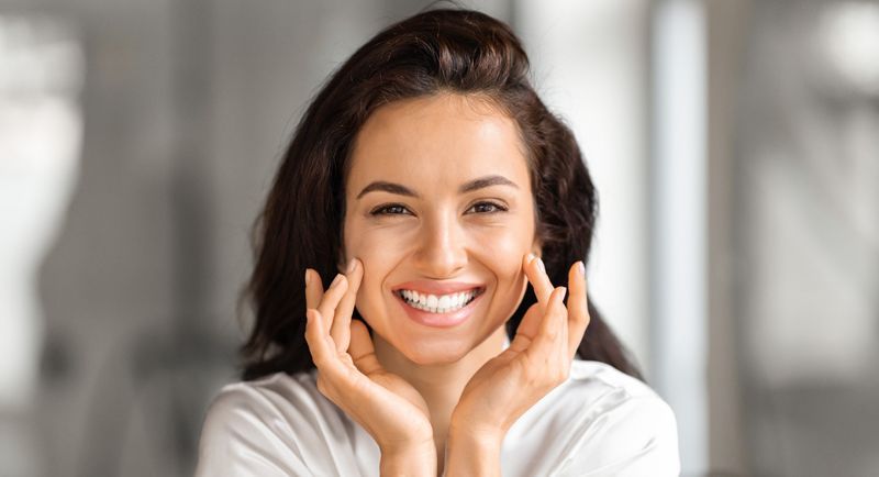 Woman with gray hair smiling in mirror, hands on her face; light pink top; neutral background.