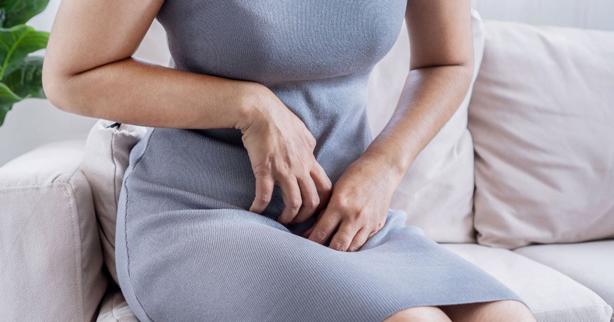 Woman in a gray dress seated on a sofa, holding her hand near her crotch, possibly experiencing discomfort, needing a yeast infection treatments NYC