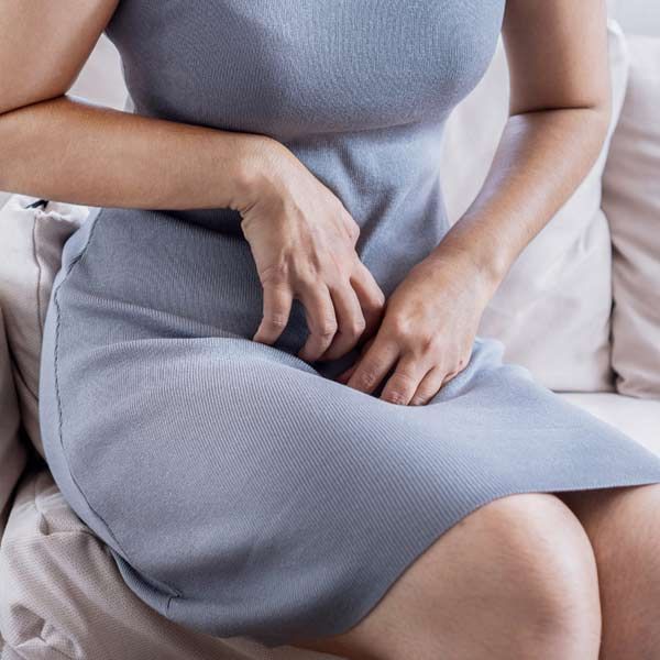 A woman in a gray dress needing yeast infection treatments is scratching her crotch, sitting on a couch.