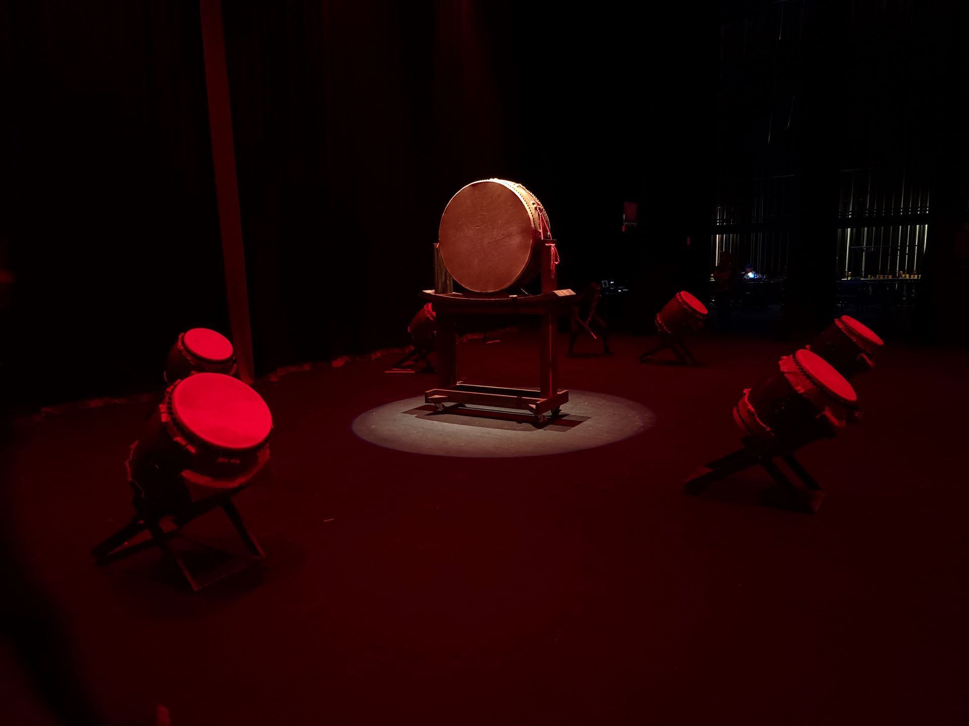 A large drum on a stand, surrounded by smaller drums. All are red-lit on a dark stage.