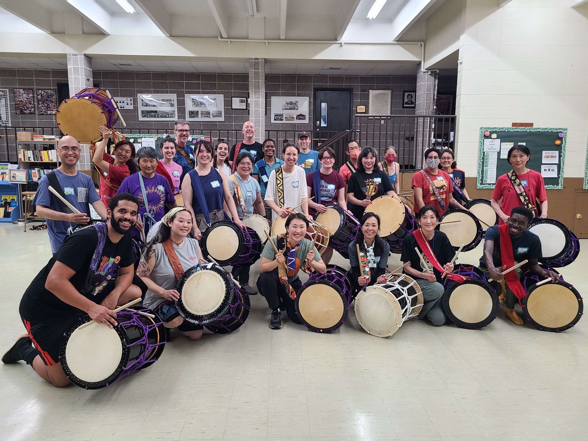 Group of people with drums indoors, posing. Some are kneeling, holding the drums. Others stand, smiling.