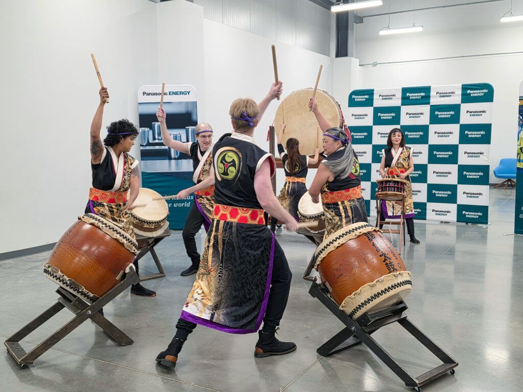 Group of people playing taiko drums in a bright indoor space. They wear traditional outfits and strike drums with sticks.