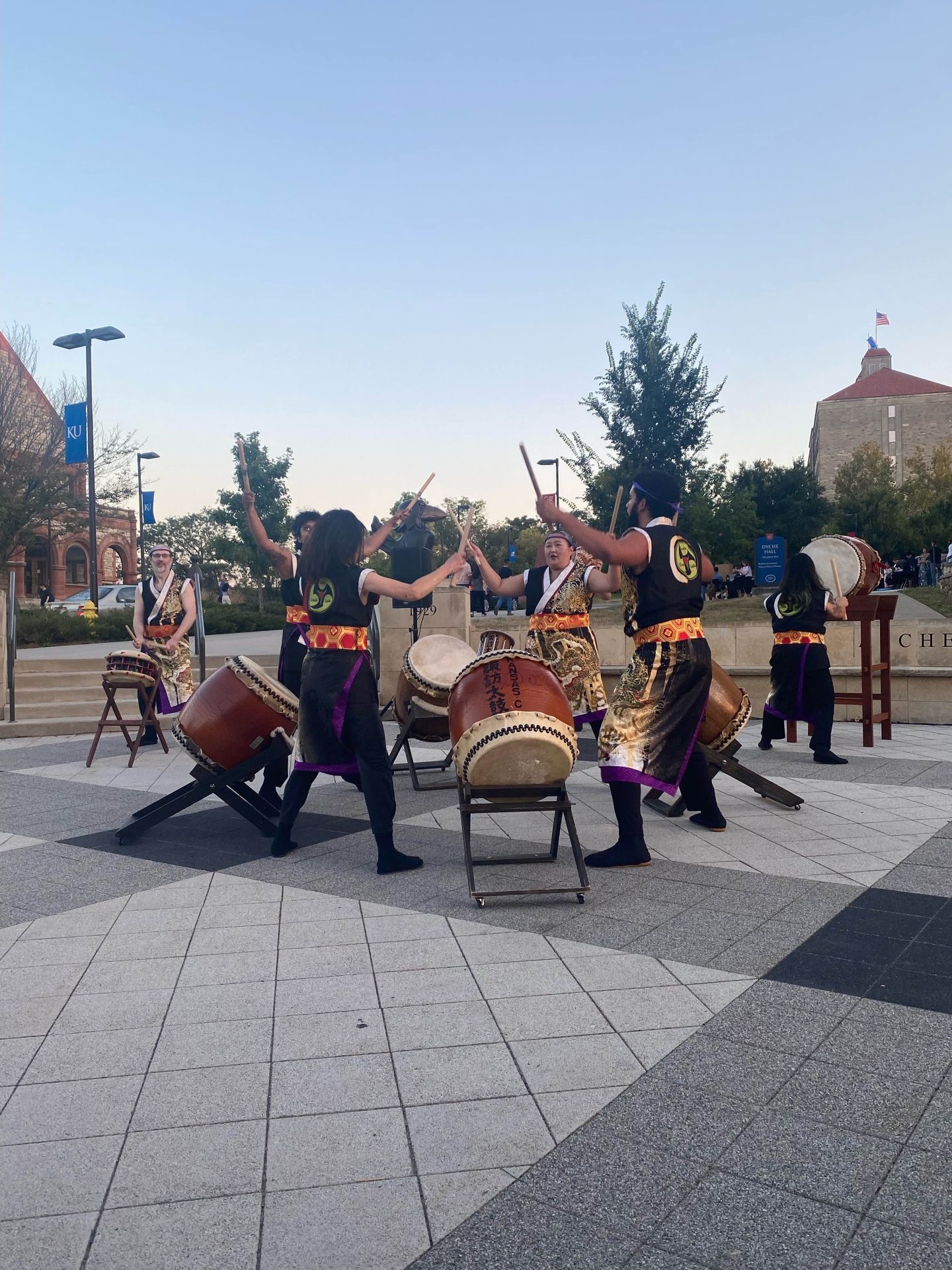 Group of people playing Taiko drums in a plaza. They are wearing black and orange attire, performing with drumsticks.