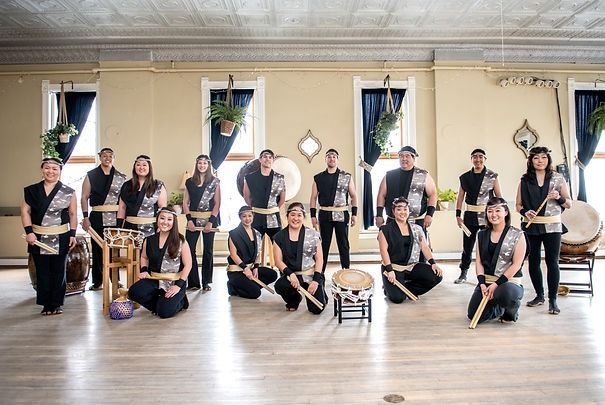 Taiko drumming group in a room, holding sticks, posing. Drums and performers dressed in black and gray.