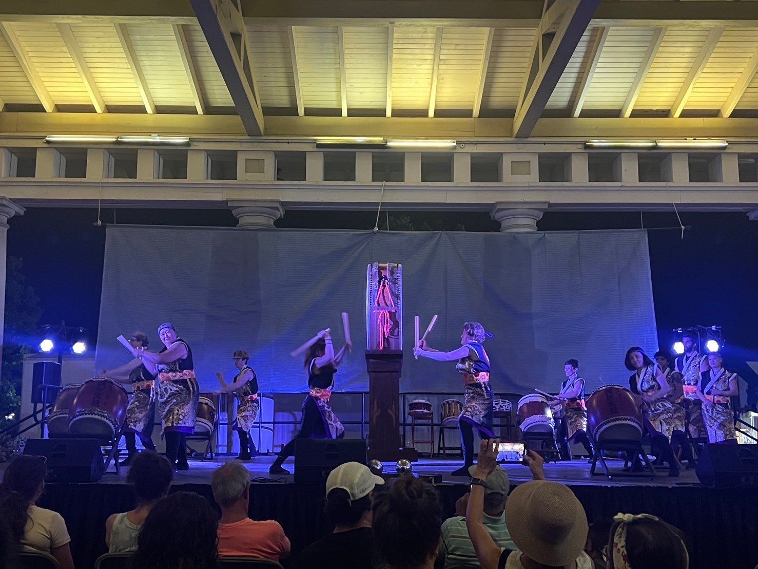 Japanese taiko drummers perform on stage, illuminated by spotlights. Audience watches.