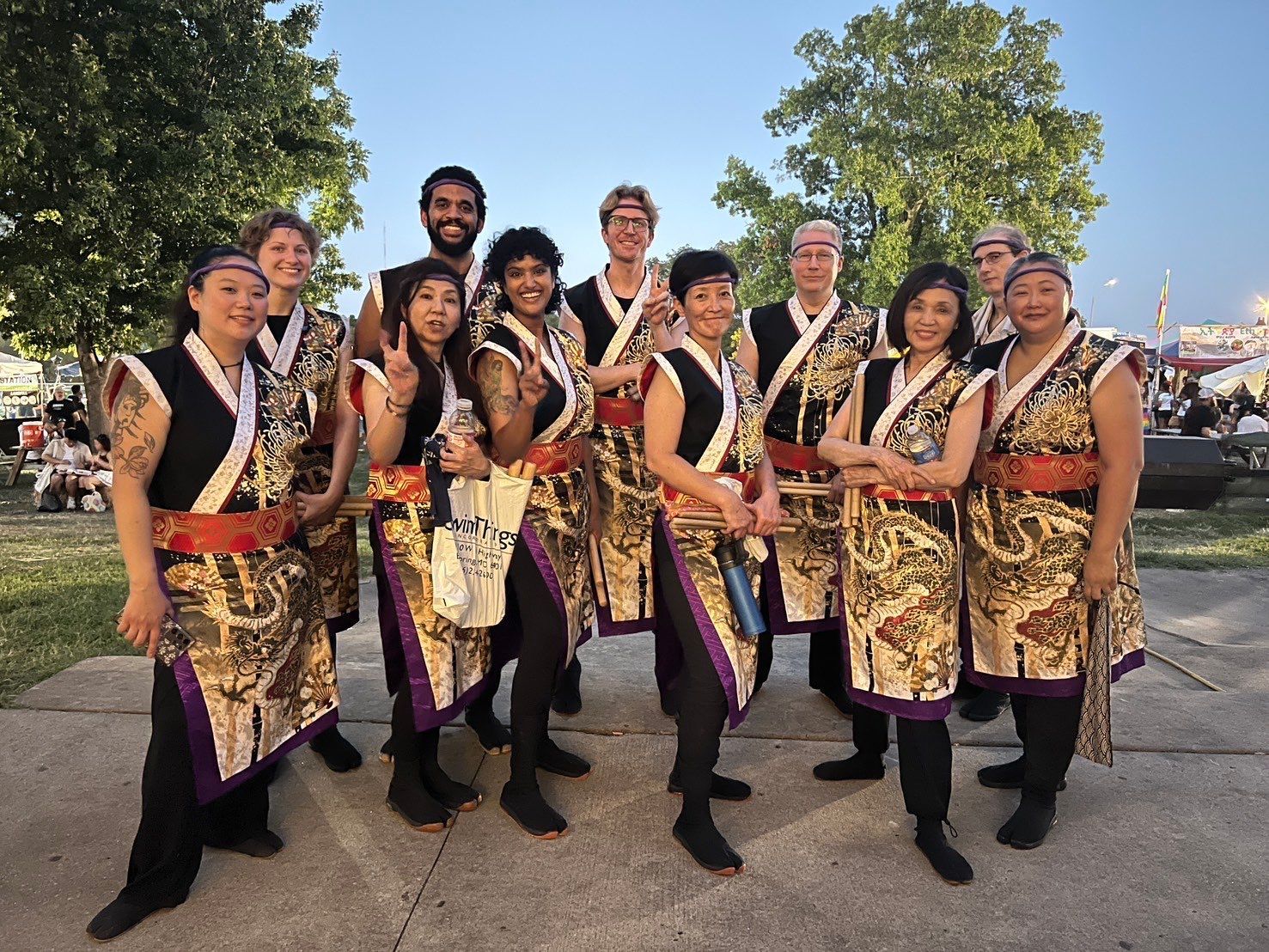 Group of performers posing in Japanese-style costumes outdoors. They smile, some hold up peace signs.