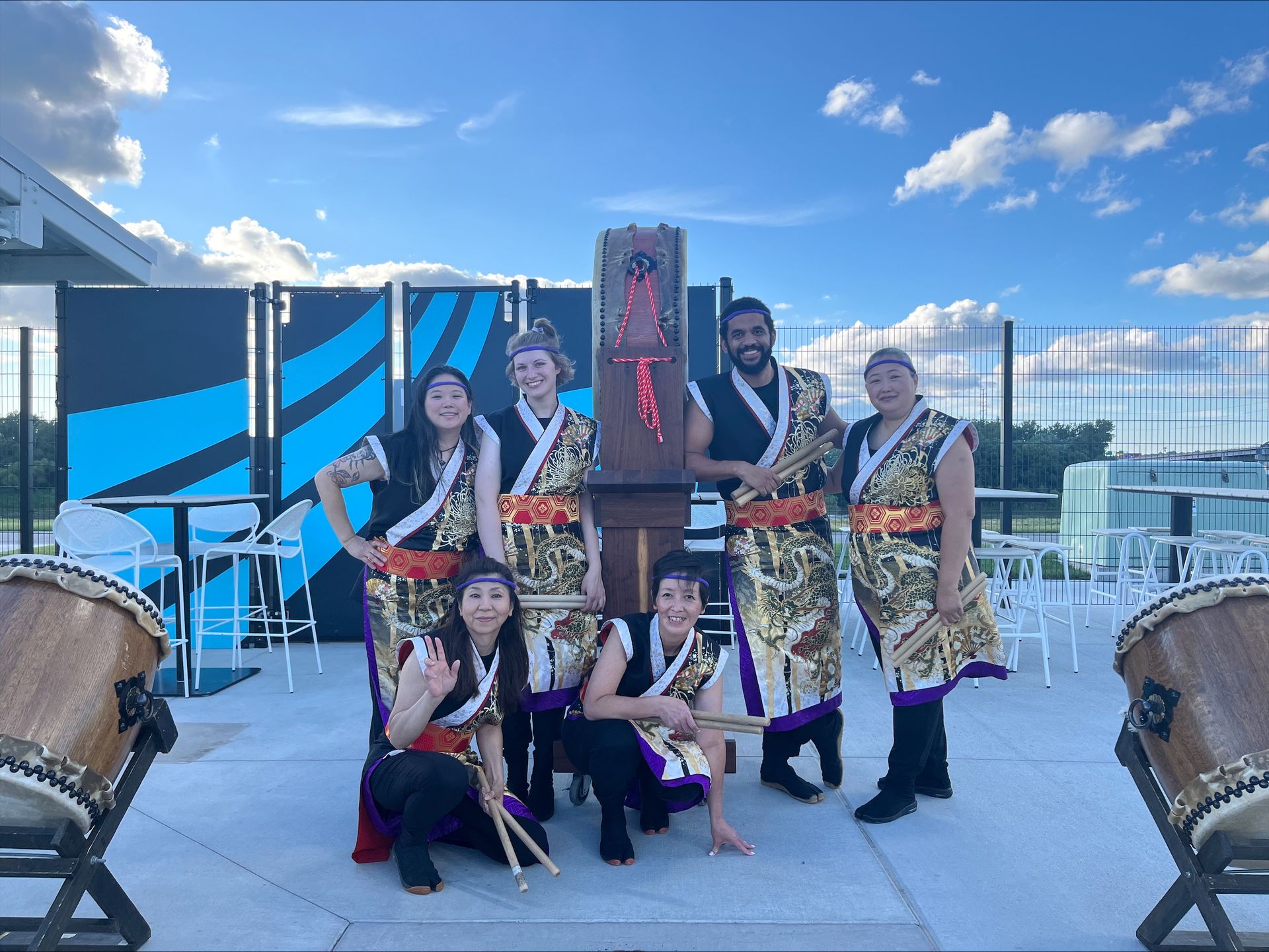 A Taiko drumming group poses outdoors. They wear traditional costumes. Blue sky, drums, and a wooden stand are visible.