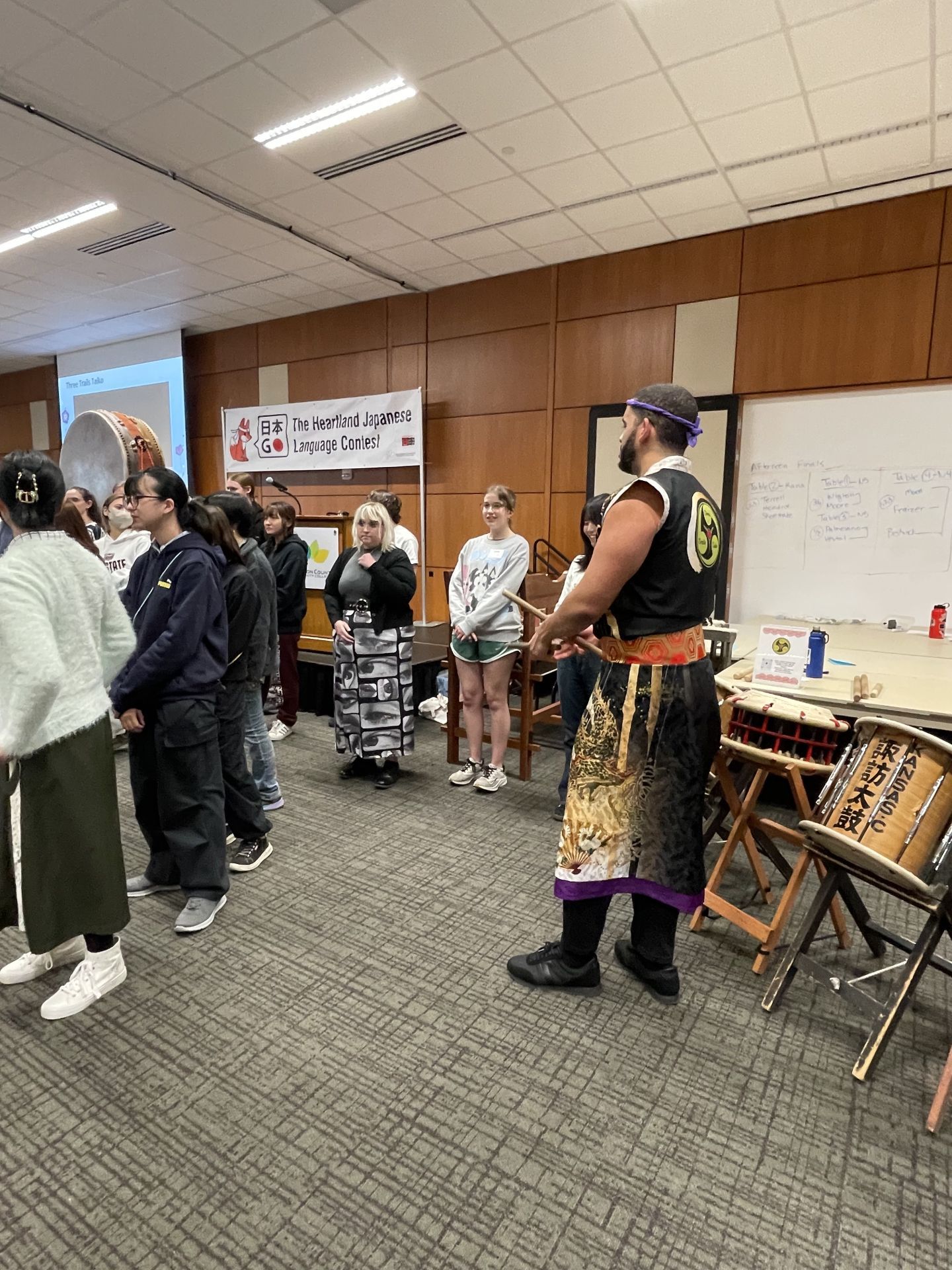 Group of people in a room, some standing near a man dressed in traditional garb.
