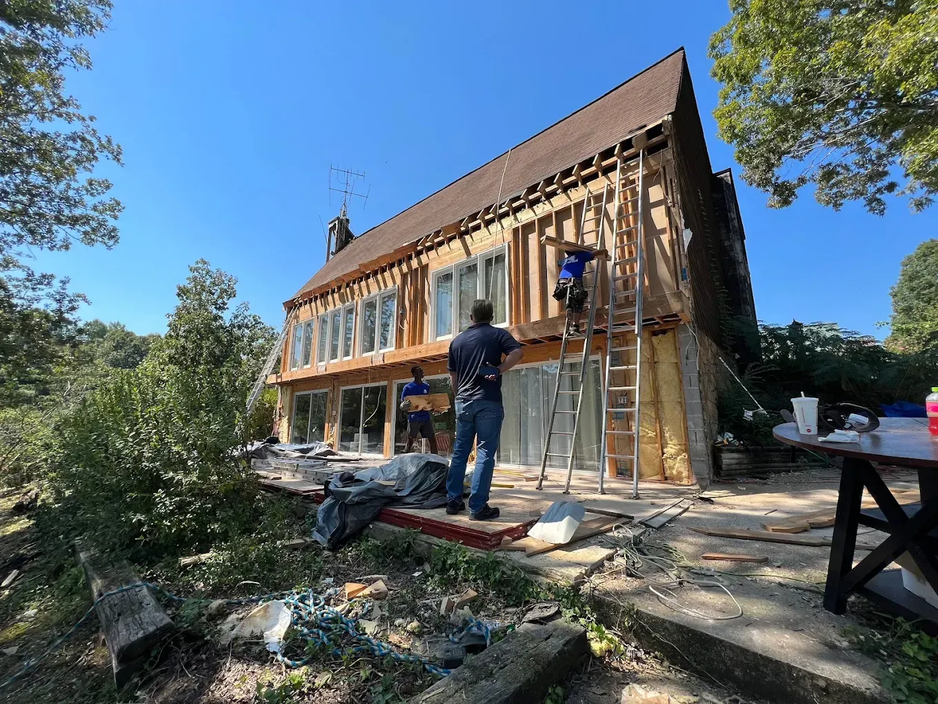 House exterior being renovated; workers on ladders, removing siding and framing. Blue sky, sunny day.