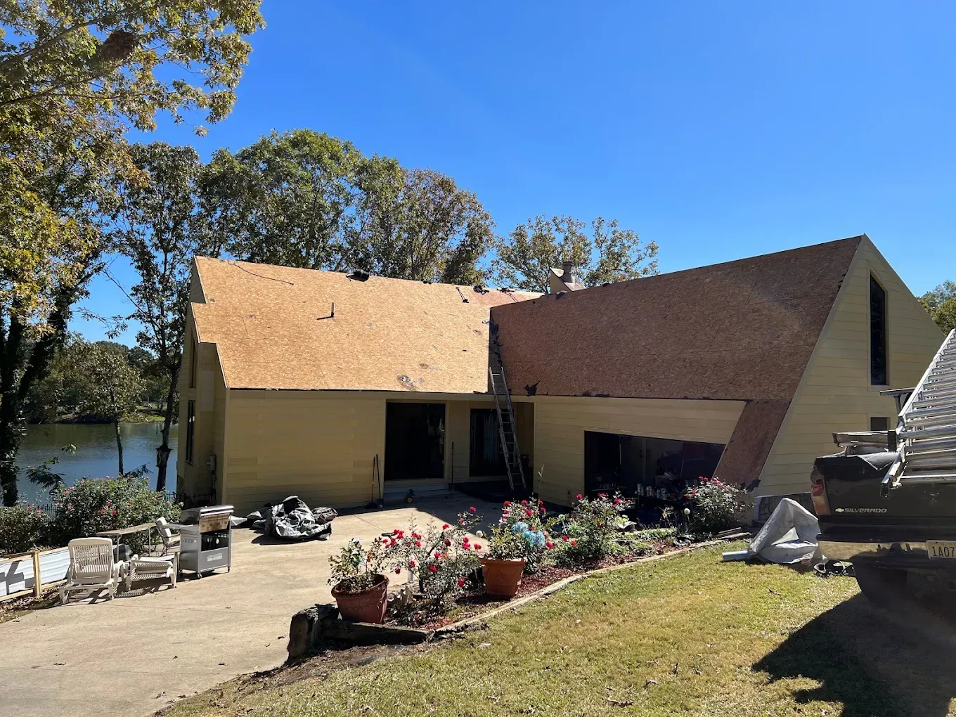 A yellow house under roof repair with exposed wood, set on a sunny day next to water and trees.