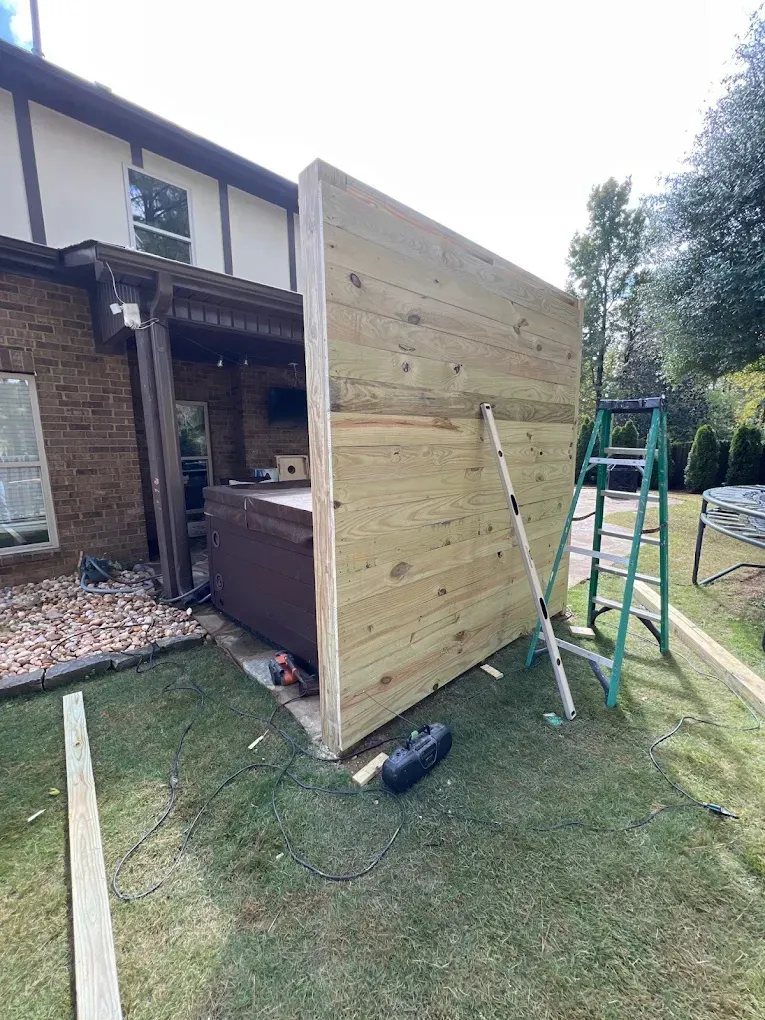Wooden privacy wall partially built beside a hot tub on a grassy lawn. A ladder and tools are present.