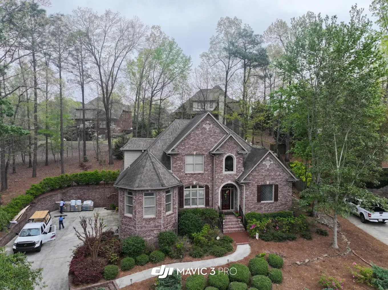 Drone view of a brick house on a hill, surrounded by trees. White trucks and people are in the driveway.