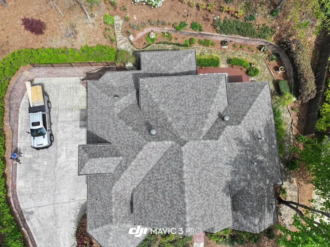 Overhead view of a house with a gray shingle roof, driveway, and a truck parked beside it, surrounded by greenery.