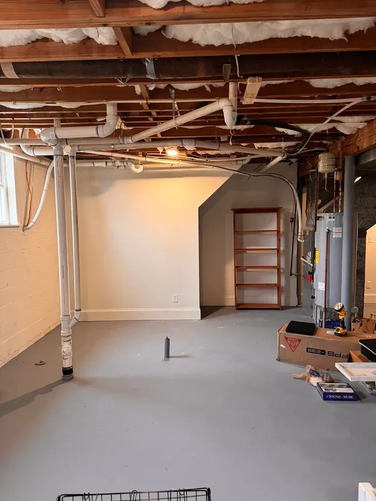 Basement interior with exposed ceiling beams, plumbing, and a gray floor. A white wall, shelves, and water heater are visible.