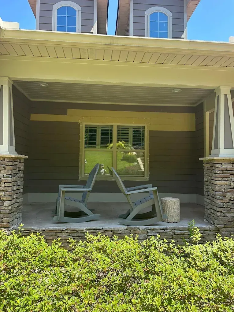 Two gray rocking chairs on a porch with stone columns and shutters; green bushes in front.