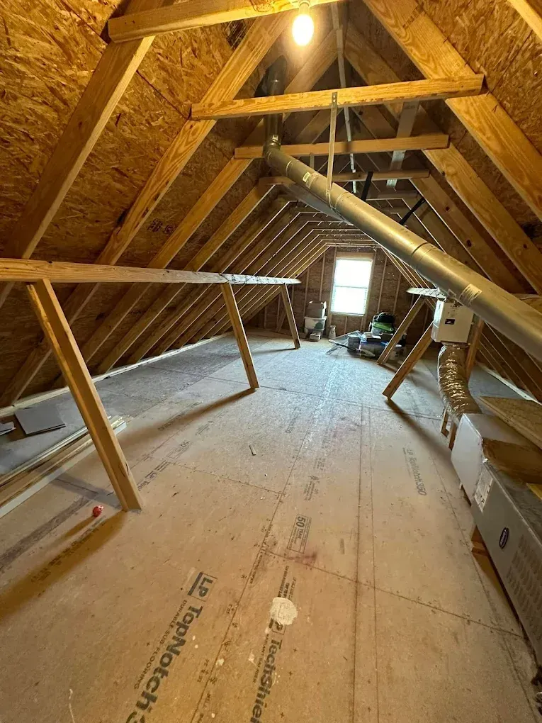 Attic interior with exposed wood beams, ductwork, and partial flooring. A light bulb hangs from the peak.