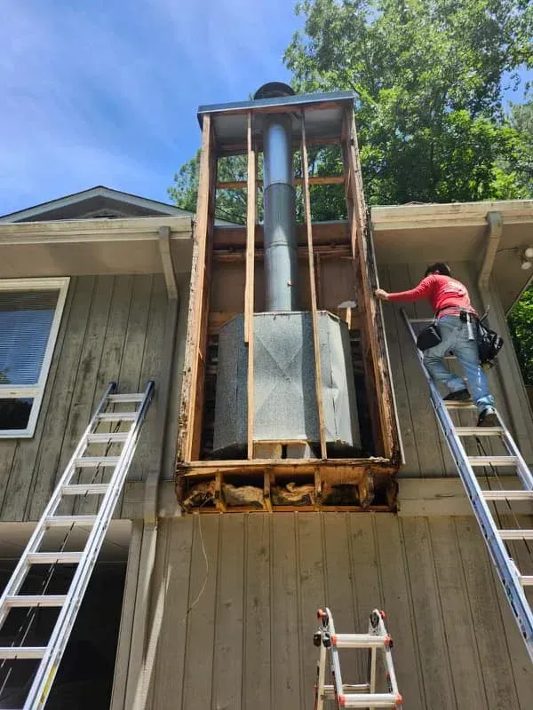 Construction worker repairing chimney exterior with ladders. Exposed frame, metal flue, and weathered siding.
