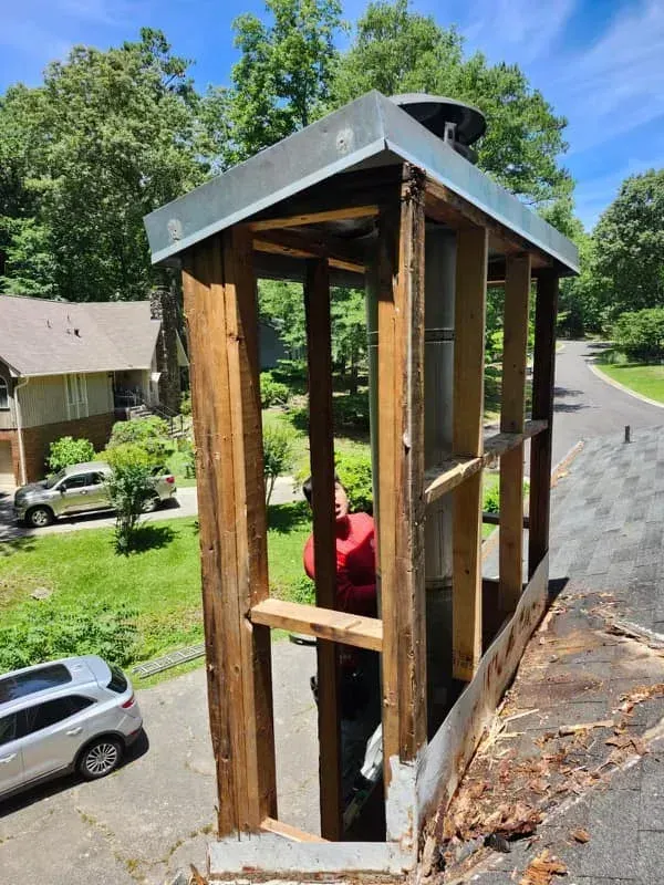 Man repairs a chimney, removing weathered wood framing. Silver car, green trees, roof, and road in the background.