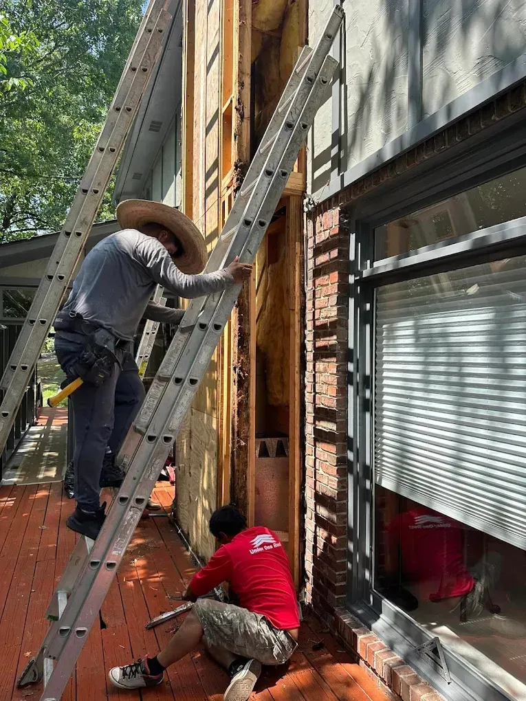 Two workers repairing exterior wall siding next to a window; one on a ladder, one kneeling.