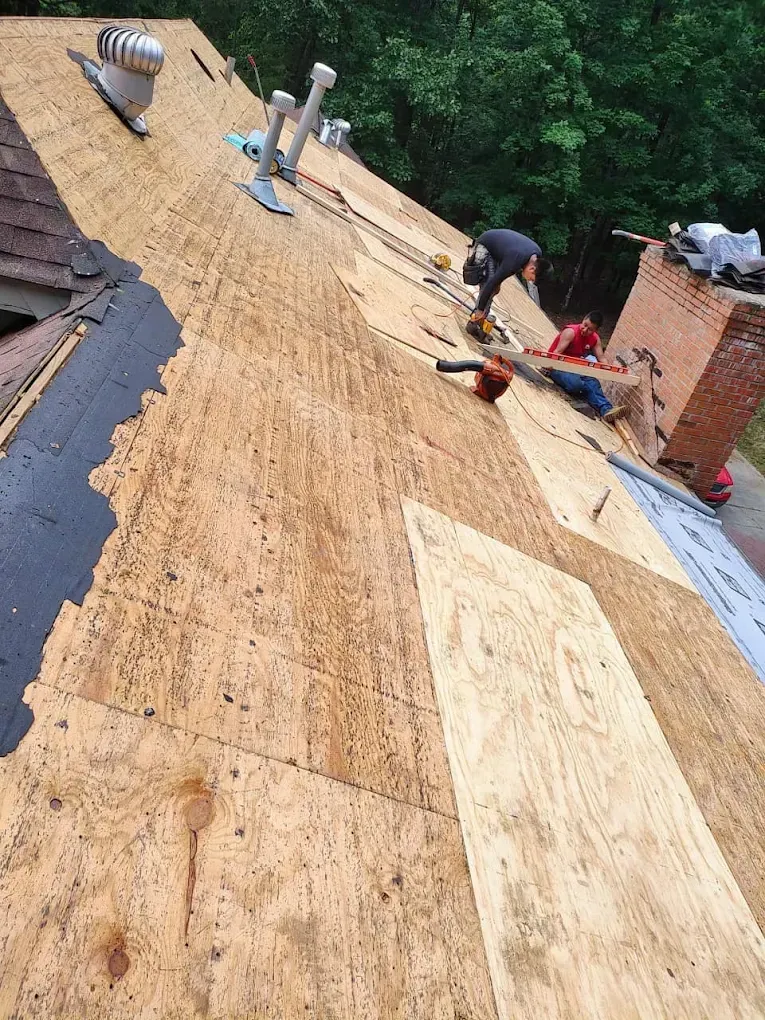Roofers working on a roof, removing old shingles and replacing plywood. Chimney and vents are visible.