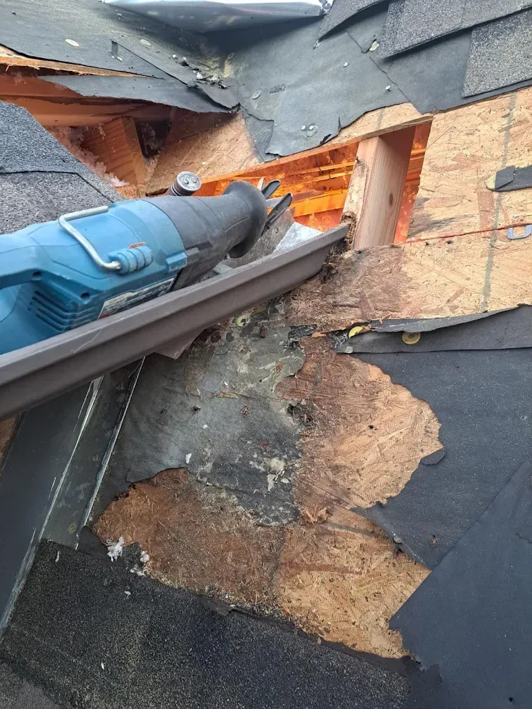 Person using a reciprocating saw on a damaged roof, cutting away wood and shingles.