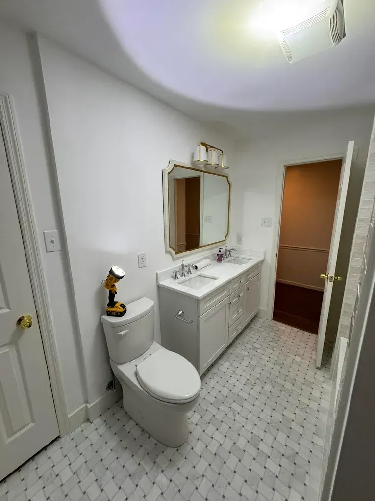 Bathroom with white walls, marble-like floor, white vanity, gold-framed mirror, toilet, and an open doorway.