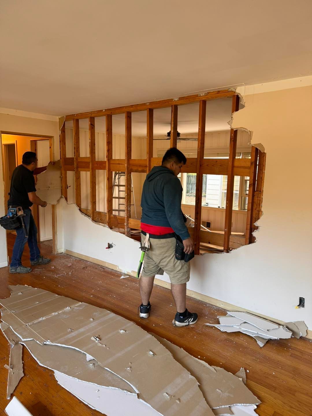 Two workers tearing down a wall during a home renovation, exposing the wooden frame. Drywall debris on the floor.
