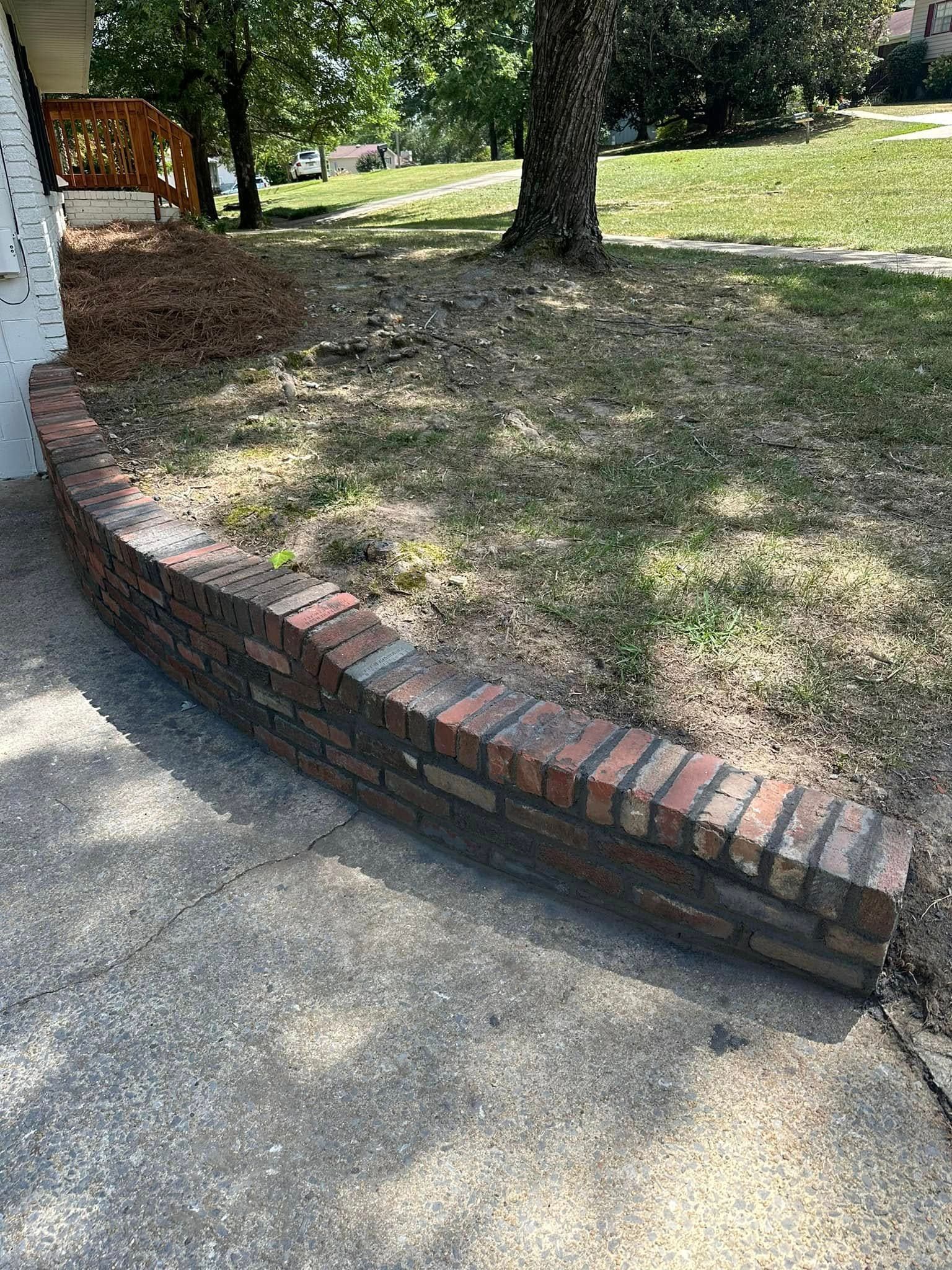 A low brick retaining wall curves along a concrete sidewalk next to a house with a grassy area.