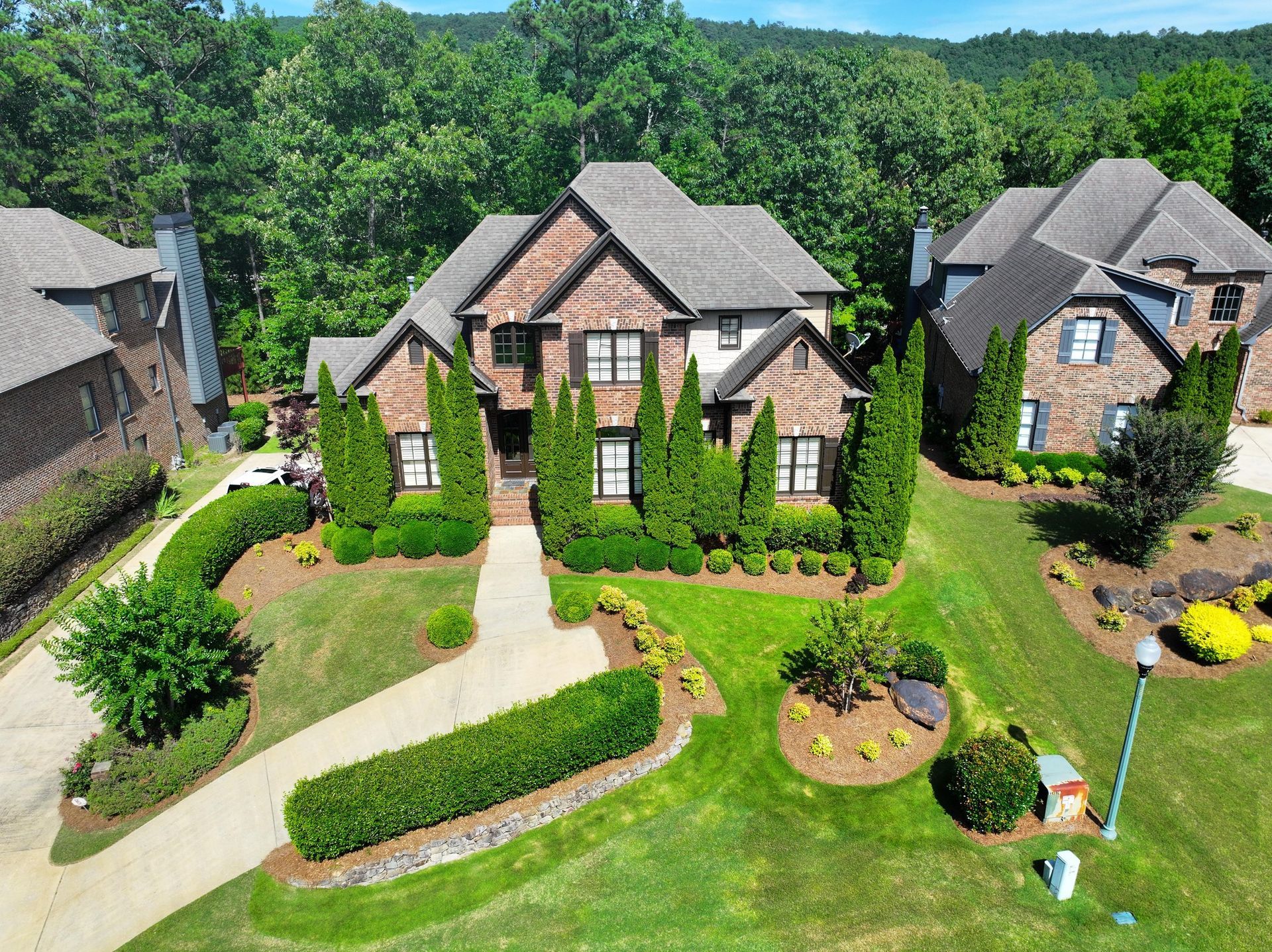 House with brown roof, green lawn, trees. Driveway curves to the entrance. Other houses visible.