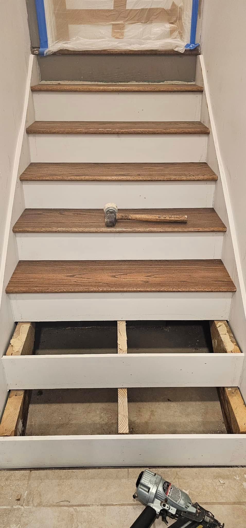 A staircase under construction; white risers, brown treads, a hammer rests on a step, and the wall at the top is taped.