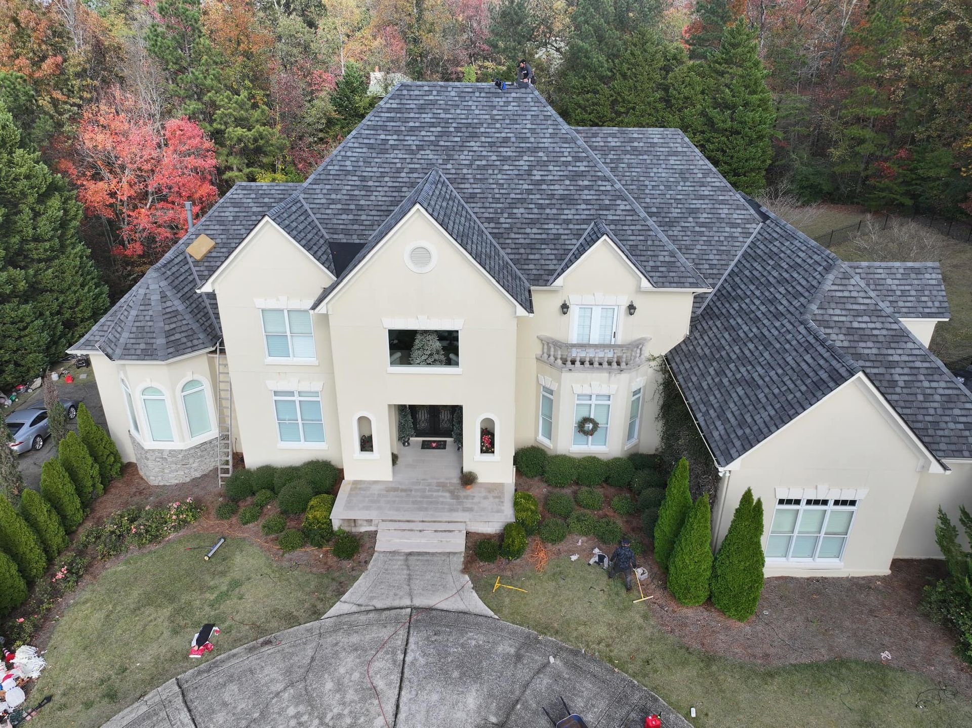 Large beige house with gray shingle roof, surrounded by trees with fall foliage and green bushes.