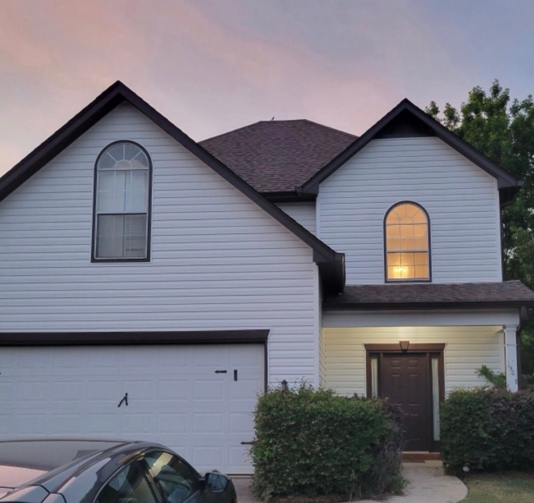 Two-story house with white siding, arched windows, and brown roof under a sunset sky.